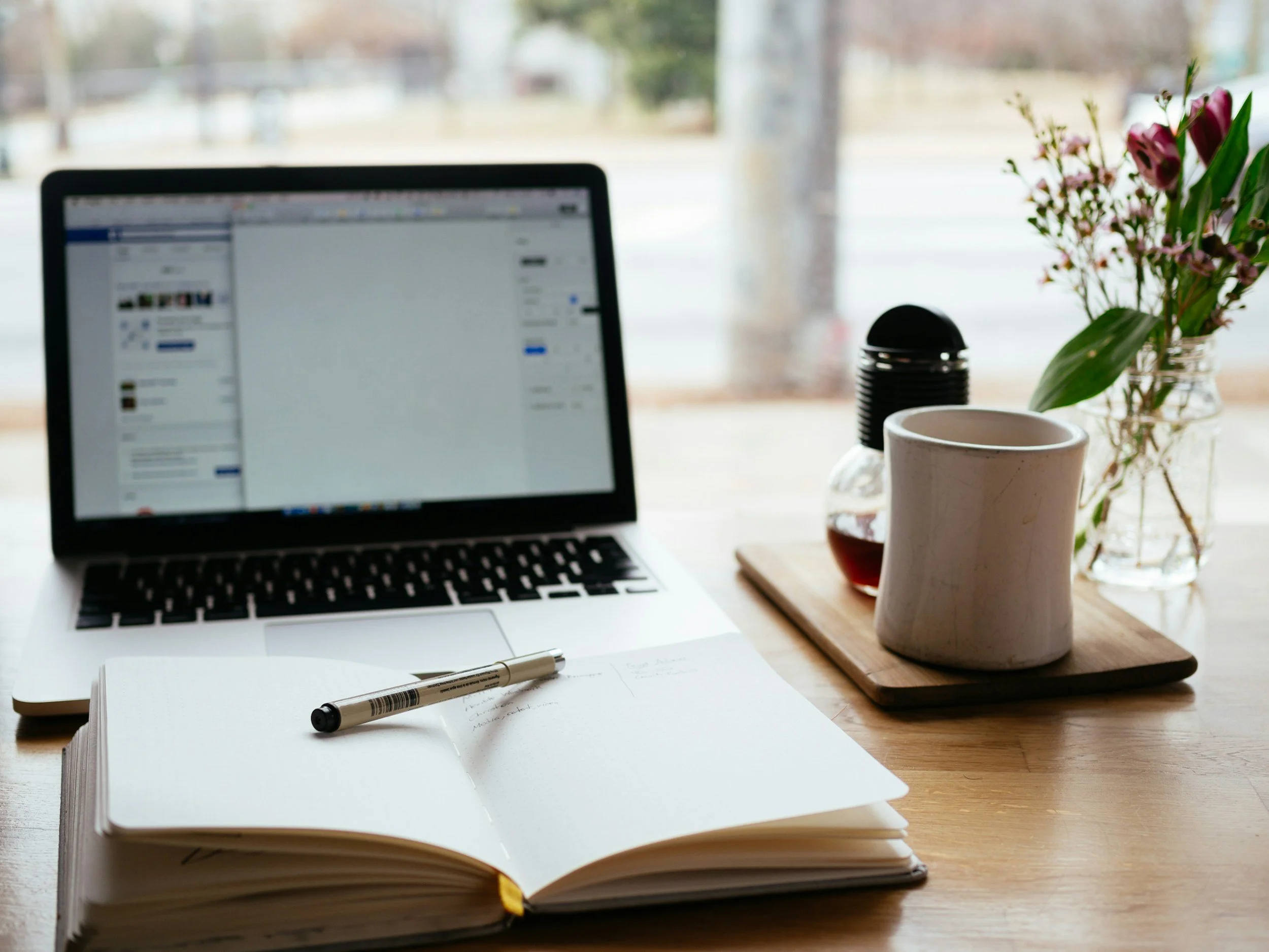 A workspace with a notebook, laptop, flowers and a mug